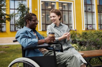 Young black man with glasses wearing long-sleeved blue shirt sitting in a wheelchair holding coffee cup talking to a smiling white woman wearing a belted gray dress showing him an image on a cell phone