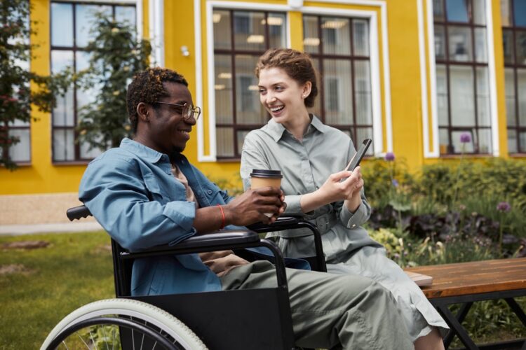 Young black man with glasses wearing long-sleeved blue shirt sitting in a wheelchair holding coffee cup talking to a smiling white woman wearing a belted gray dress showing him an image on a cell phone